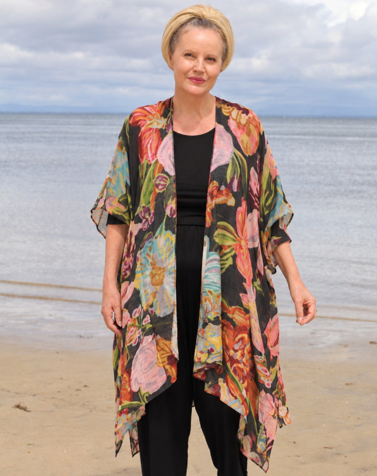 A woman standing on a beach wearing a black kimono-style cardigan with a vibrant multicolor floral print, layered over a black top and black pants.