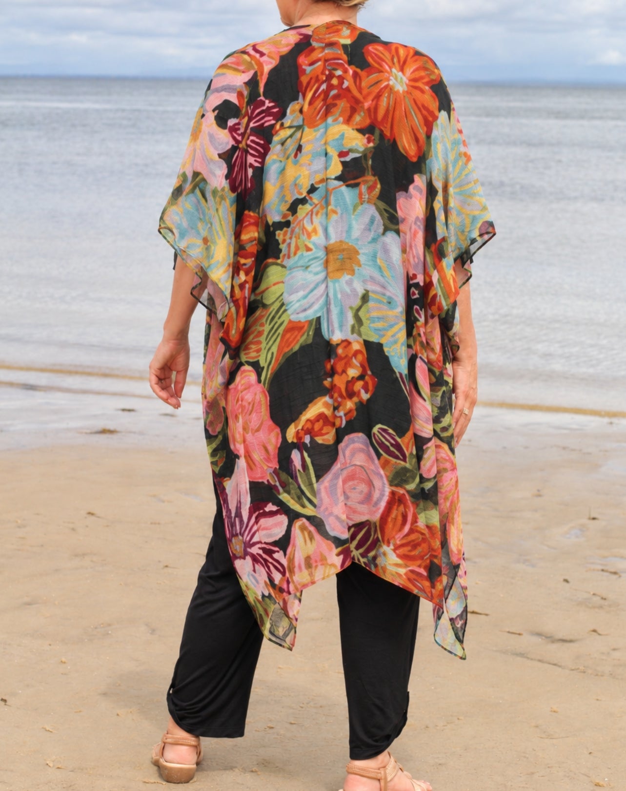 A woman standing on a sandy beach facing away from the camera, wearing a loose black floral kimono-style top with large multicolored flowers and black pants.