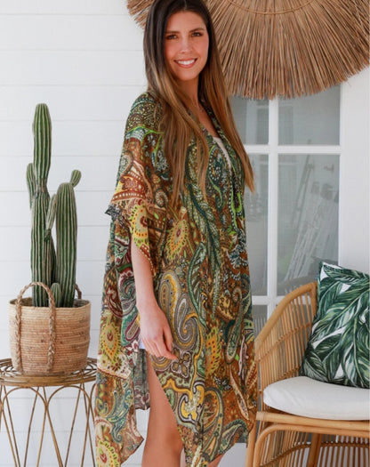Woman wearing a paisley print in green kimono cover up jacket standing in a room with a boho wicker chair.
