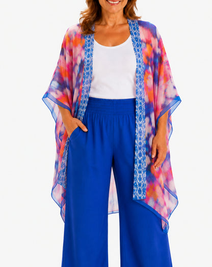 A woman wearing a lightweight open-front kimono cardigan with a vibrant blue, pink, and orange tie-dye pattern and blue patterned trim, styled over a white tank top and bright blue wide-leg pants.