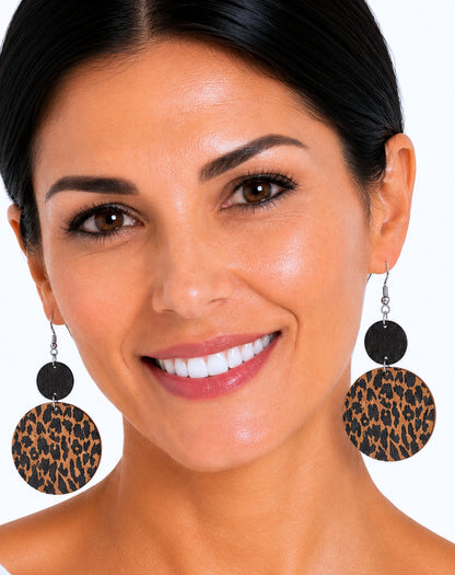 A close-up of a smiling woman wearing large drop earrings with black circular discs and leopard-print circular discs below, against a plain white background.