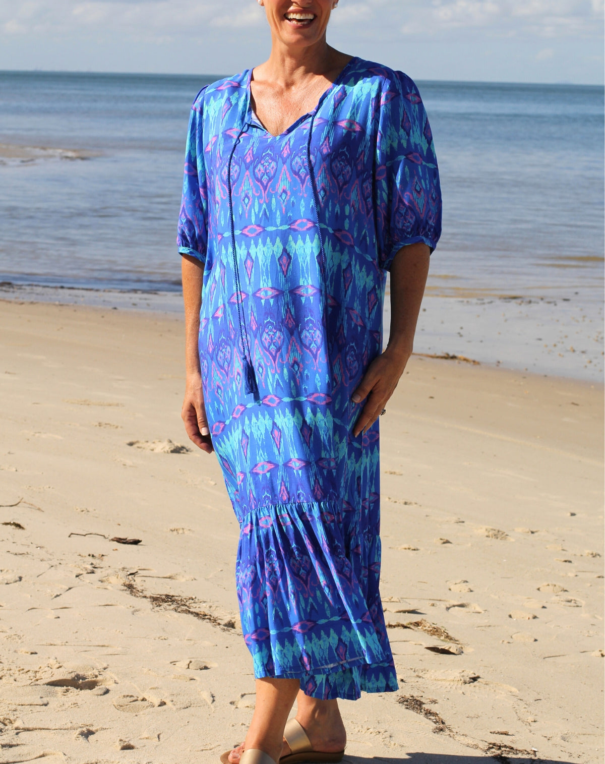 A woman wearing a blue and purple patterned ikat boho midi dress stands on a sandy beach with the ocean in the background.