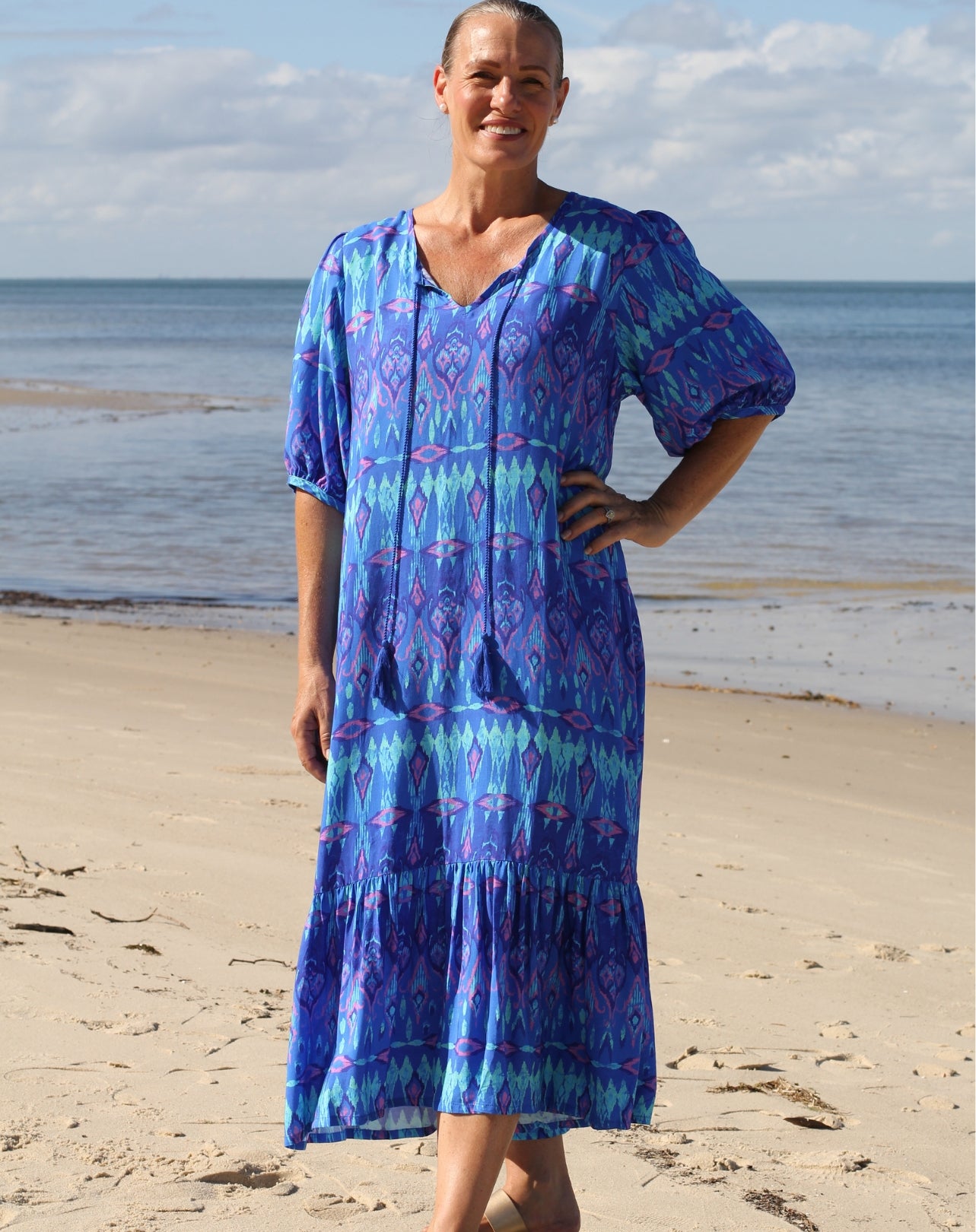 A mid-sized woman wearing a blue and purple ikat boho beach dress stands on a sandy beach with the ocean and cloudy sky in the background.