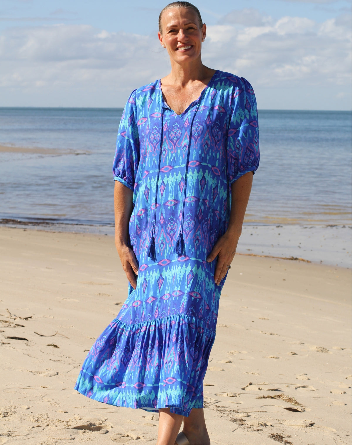 A woman wearing a blue and purple ikat patterned boho midi dress stands on a sandy beach with the ocean and sky in the background