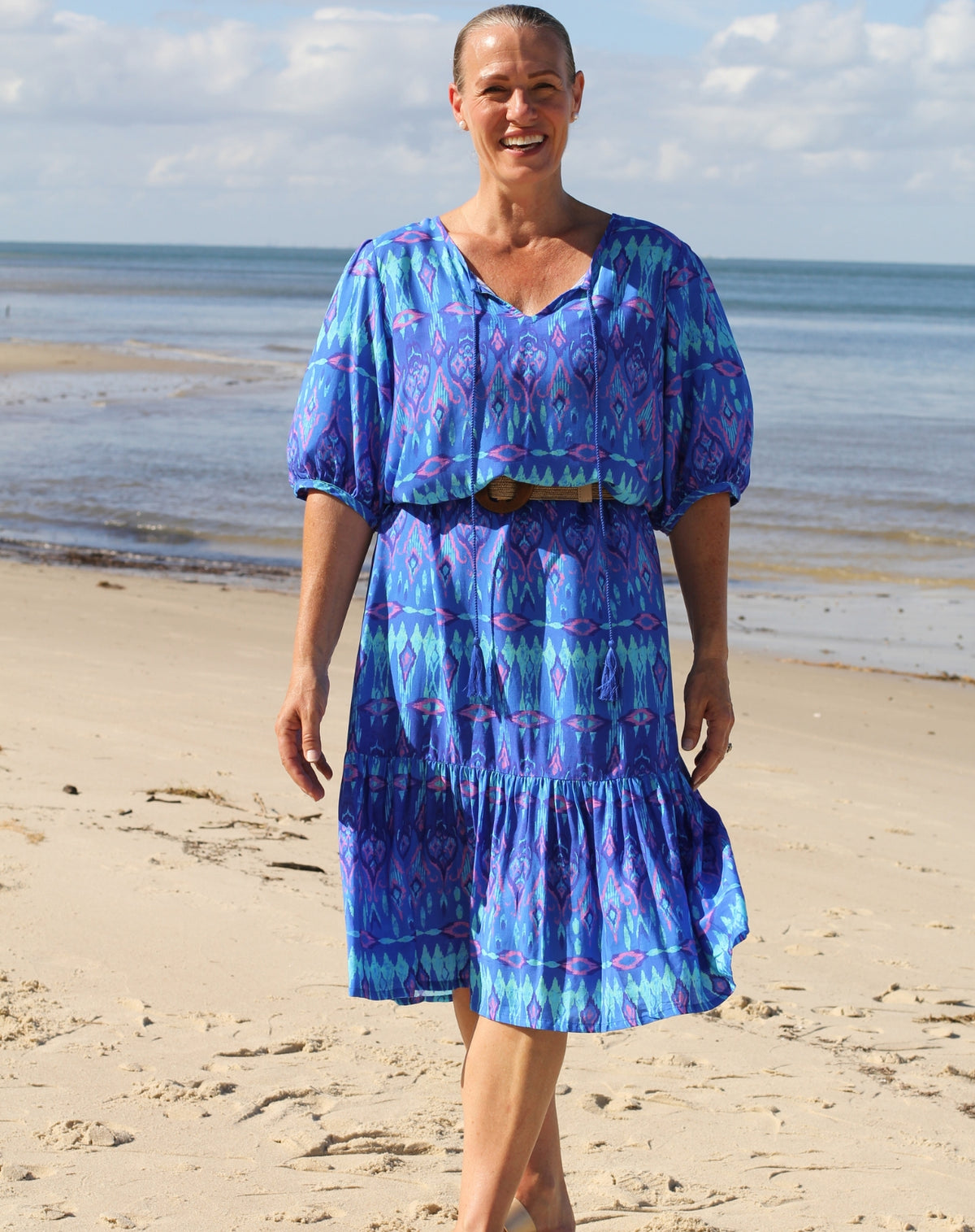 A woman stands on a sandy beach wearing a blue patterned midi dress with short puff sleeves and a V-neckline. The dress has an ikat-style print in shades of blue, purple, and pink, and is paired with a brown belt.