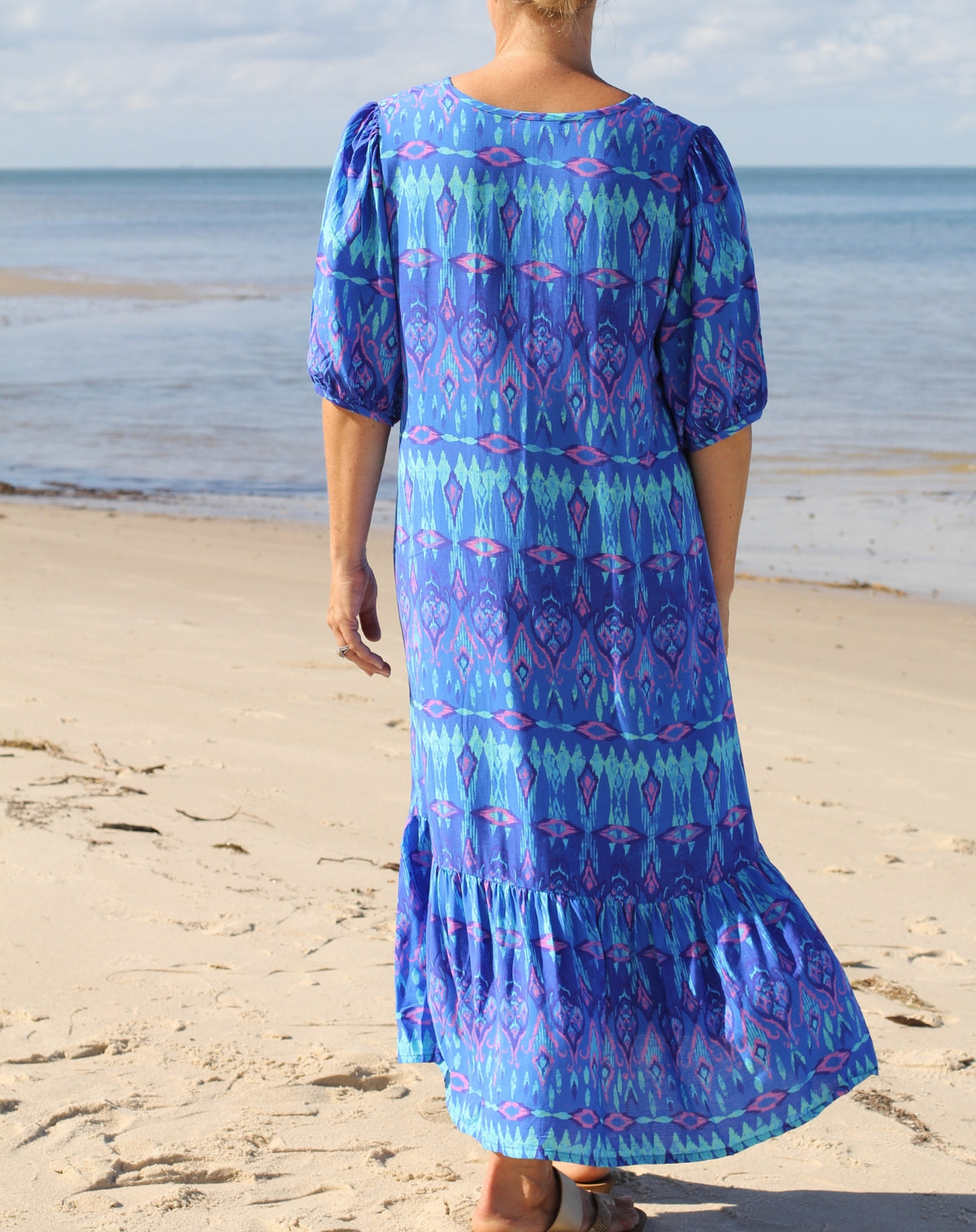 A woman viewed from behind wearing a blue and purple ikat print maxi dress with puff sleeves, standing on a sandy beach with the ocean in the background.