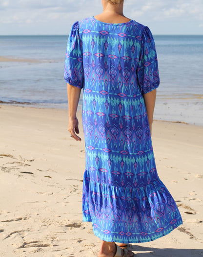 A woman viewed from behind wearing a blue and purple ikat print maxi dress with puff sleeves, standing on a sandy beach with the ocean in the background.