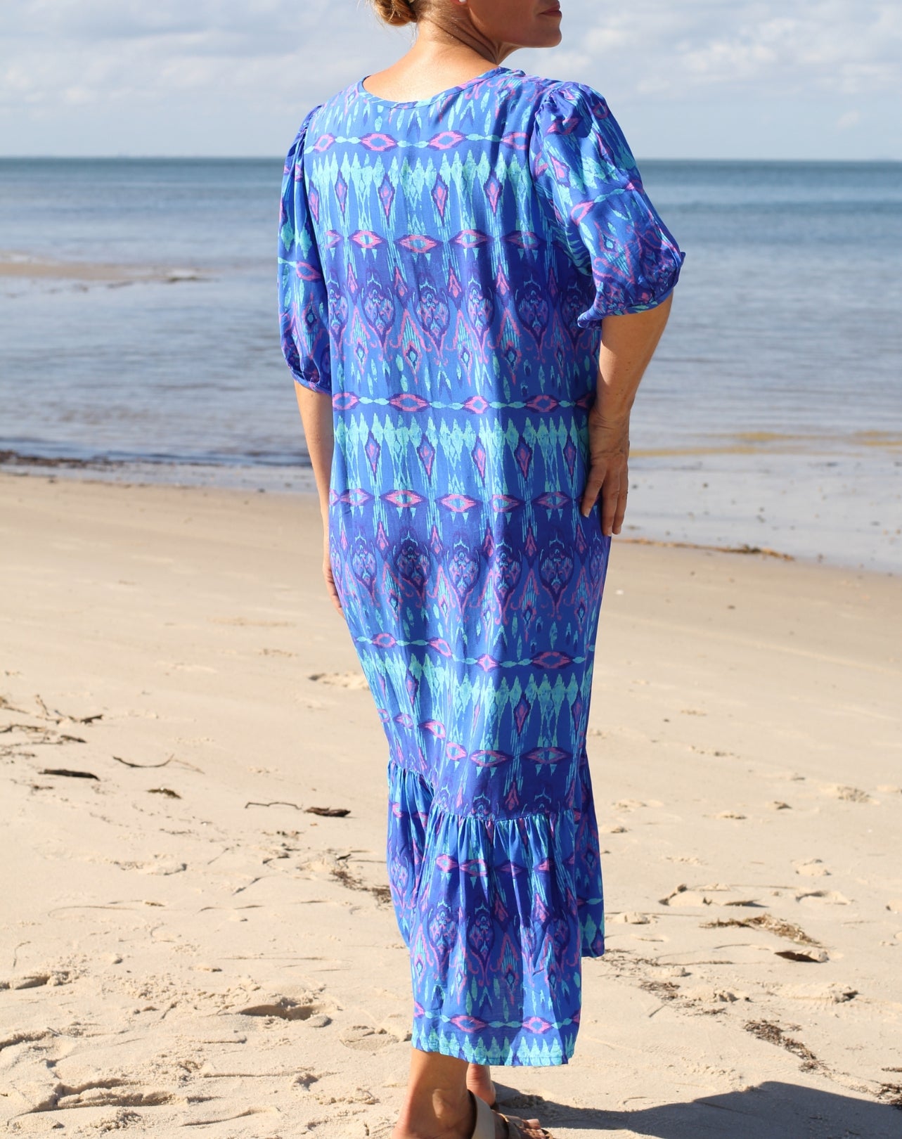 A woman stands on a sandy beach wearing a blue patterned maxi dress with short sleeves and a ruffled hem, photographed from the back.