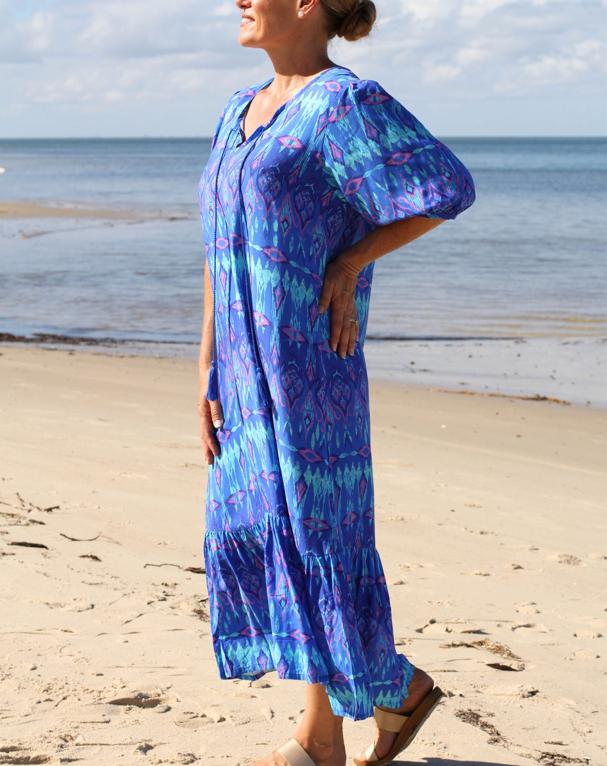 A woman wearing a blue and purple patterned midi maxi boho dress stands on a sandy beach with the ocean and cloudy sky in the background.