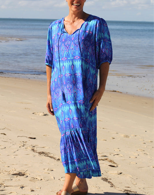 A woman wearing a blue and purple ikat patterned boho dress stands on a sandy beach with the ocean in the background.