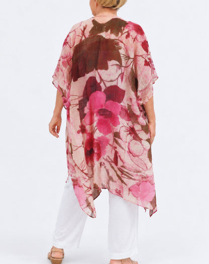 A woman shown from the back wearing a loose, flowy floral kimono top with wide short sleeves. The top has a pink, red, and brown floral pattern on a light pink base, paired with white pants and tan sandals, against a plain white studio background.
