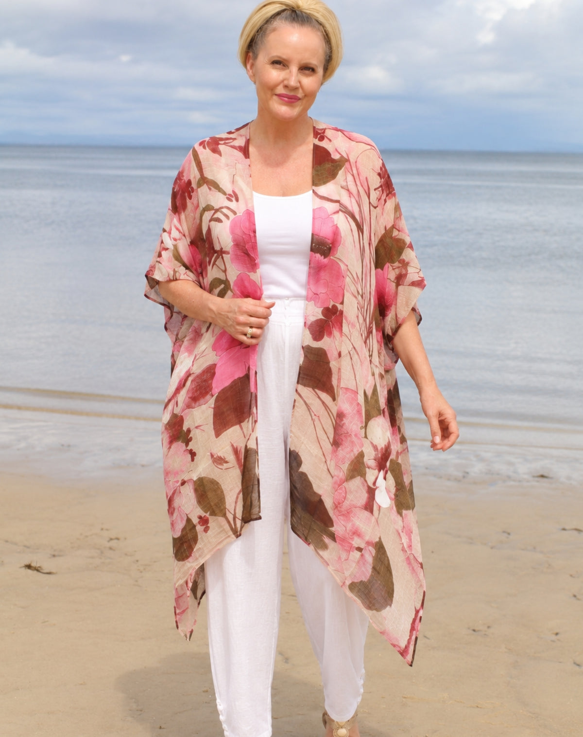 A woman standing on a sandy beach wearing a lightweight floral kimono-style cover-up with pink blossom and brown flowers over a white outfit.