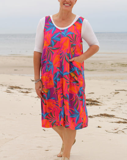 A woman standing on a sandy beach wearing a sleeveless pinafore, knee-length dress with a bold tropical floral pattern in pink, orange, and blue over a white short-sleeve top.