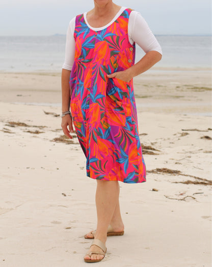 A woman wearing a sleeveless smock, knee-length dress with a vibrant pink, orange, and blue tropical floral print over a white short-sleeve top, standing on a sandy beach.