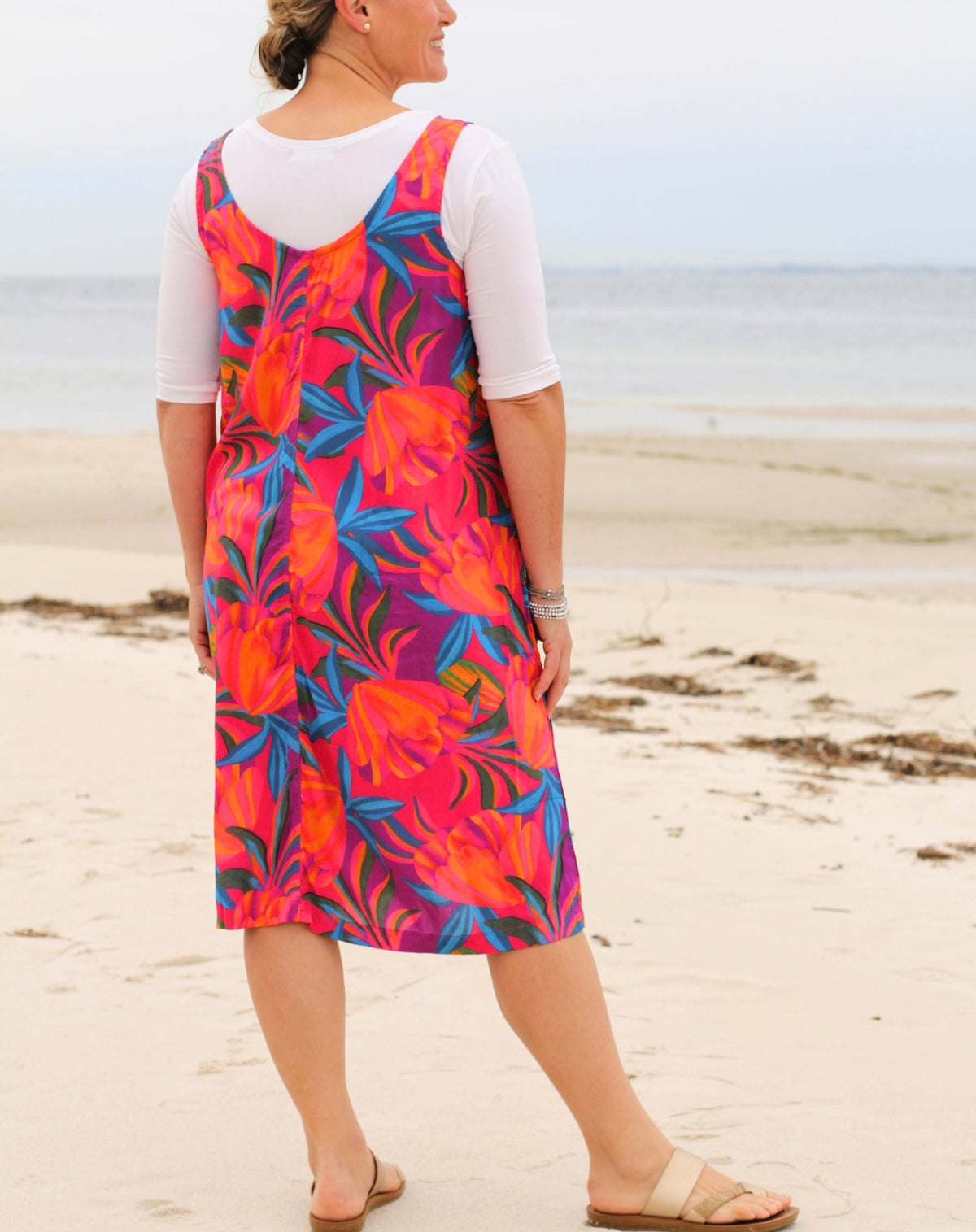 A woman standing on a sandy beach wearing a sleeveless, vibrant tropical floral dress over a white short-sleeve top. The dress features a bold pattern of orange, pink, and blue leaves.