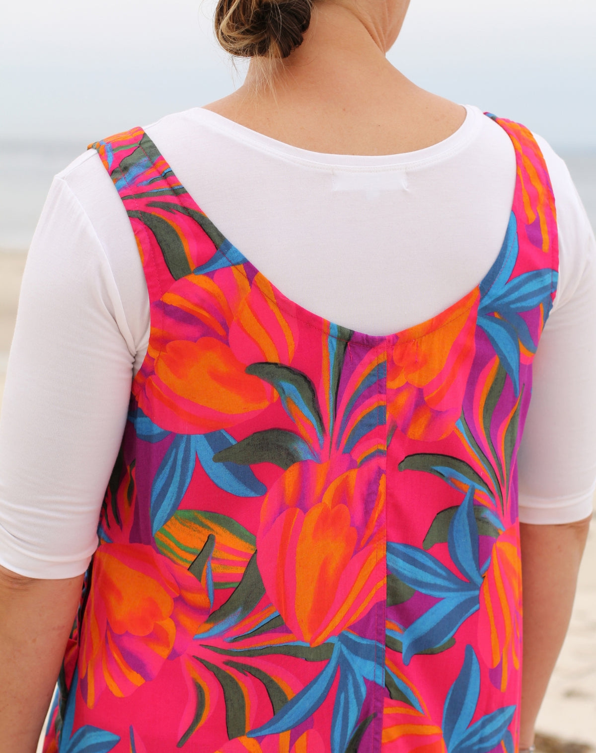 A woman viewed from behind wearing a vibrant tropical floral print smocked dress over a white short-sleeve t-shirt, set against a soft-focus beach background.