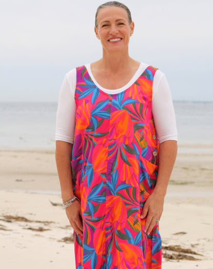 A woman standing on a sandy beach wearing a sleeveless smock dress with pockets, colorful tropical print dress over a white short-sleeve shirt.