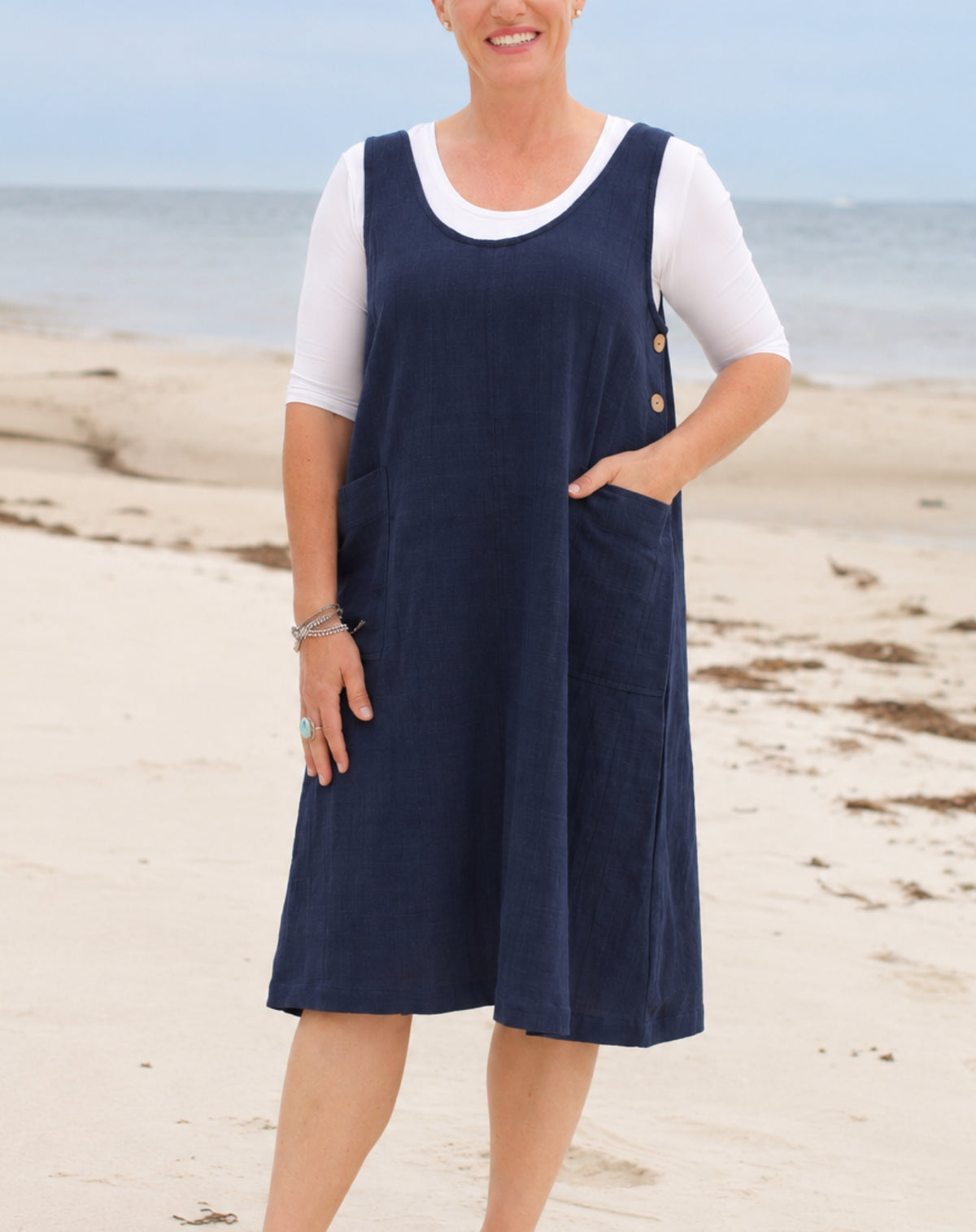 A woman standing on a sandy beach wearing a navy blue linen look pinafore dress over a white short-sleeve t-shirt.