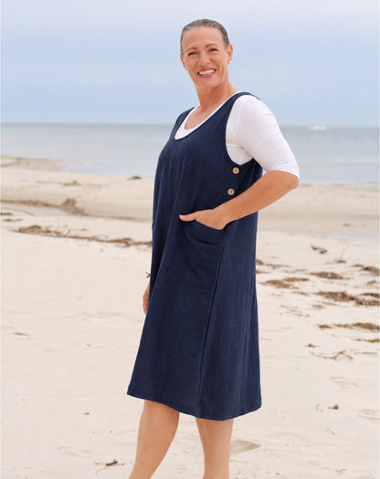 A woman standing on a sandy beach wearing a navy blue sleeveless pinafore dress over a white short-sleeve top.
