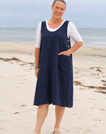 A woman standing on a sandy beach wearing a navy blue linen look pinafore dress over a white short-sleeve top.