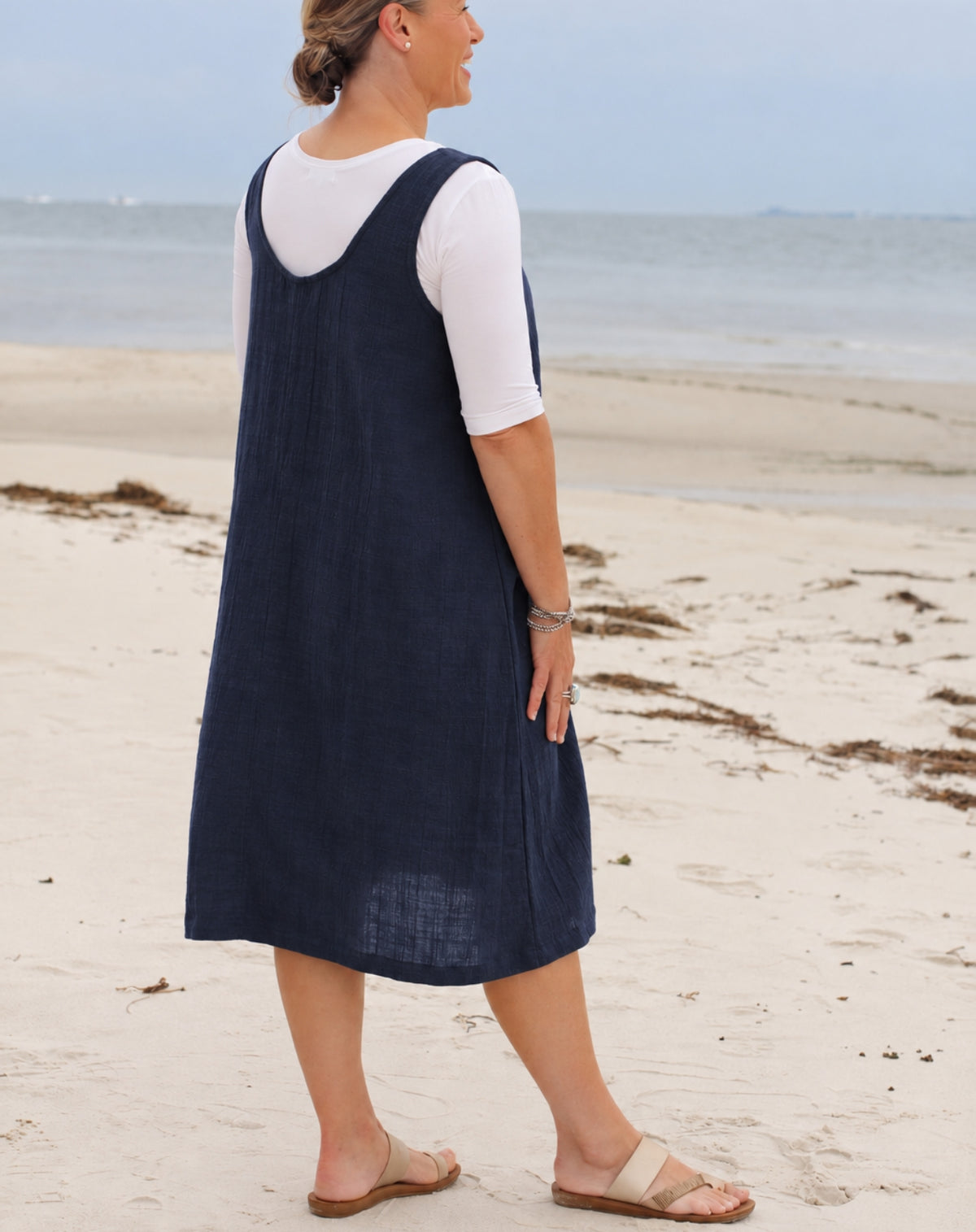 A woman standing on a sandy beach wearing a navy blue sleeveless linen pinafore dress over a white short-sleeve t-shirt, paired with beige sandals.