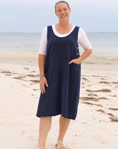 A woman standing on a sandy beach wearing a navy blue corduroy pinafore dress over a white short-sleeve top.