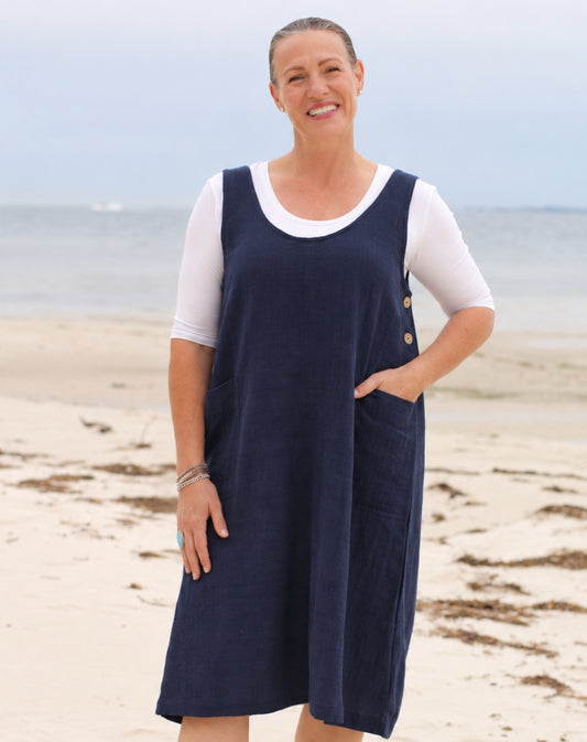 A woman standing on a sandy beach wearing a navy blue linen pinafore smock dress over a white short-sleeve top.