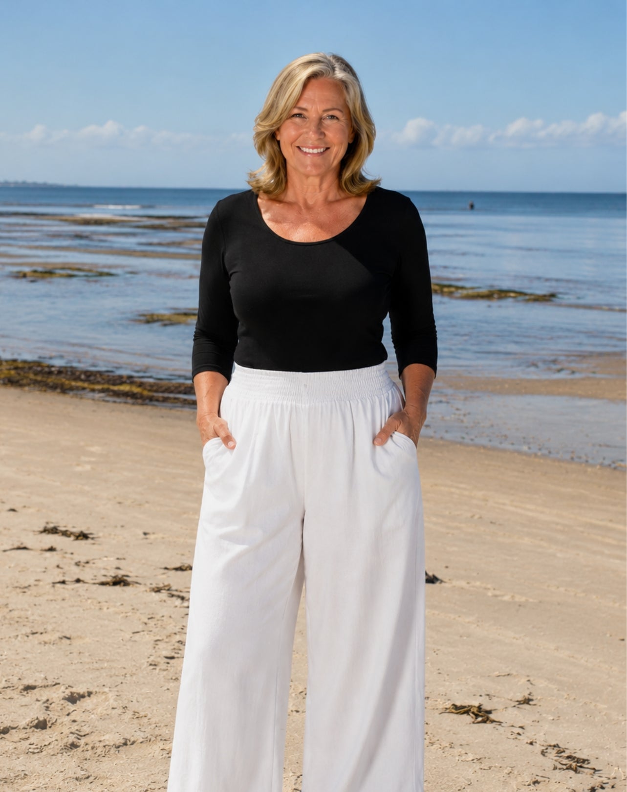 A smiling blonde woman stands on a sandy beach wearing a black three-quarter-sleeve scoop-neck top and high-waisted white wide-leg pants with side pockets.