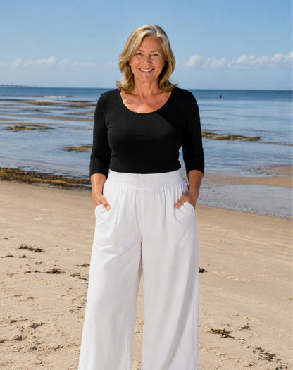 A smiling blonde woman stands on a sandy beach wearing a black three-quarter-sleeve scoop-neck top and high-waisted white wide-leg pants with side pockets.