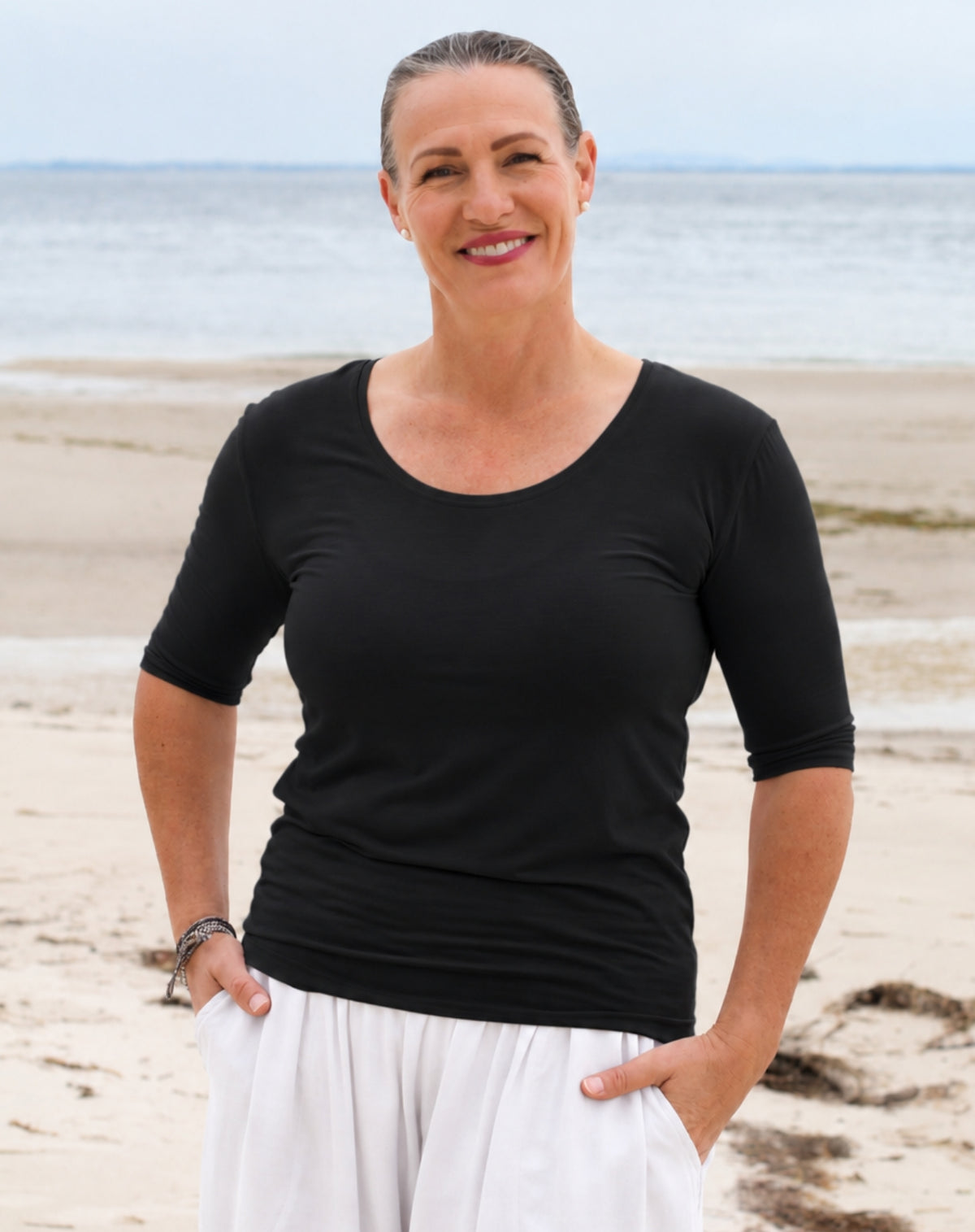 A smiling woman standing on a sandy beach with the ocean in the background, wearing a black three-quarter sleeve scoop-neck top and white pants.