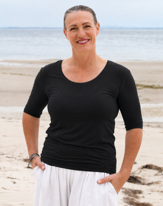 A smiling woman standing on a sandy beach with the ocean in the background, wearing a black three-quarter sleeve scoop-neck top and white pants.