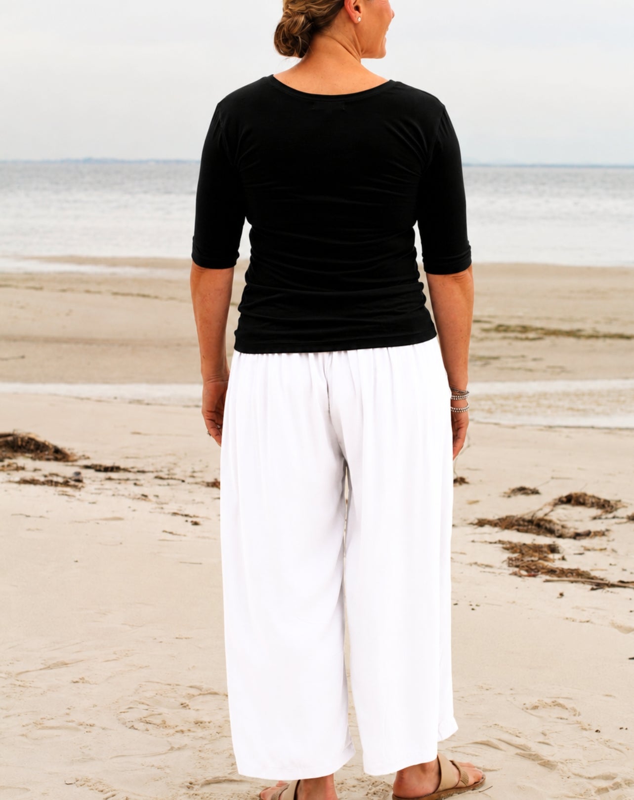 A woman standing on a sandy beach, viewed from behind, wearing a black three-quarter sleeve top and loose white wide-leg pants.