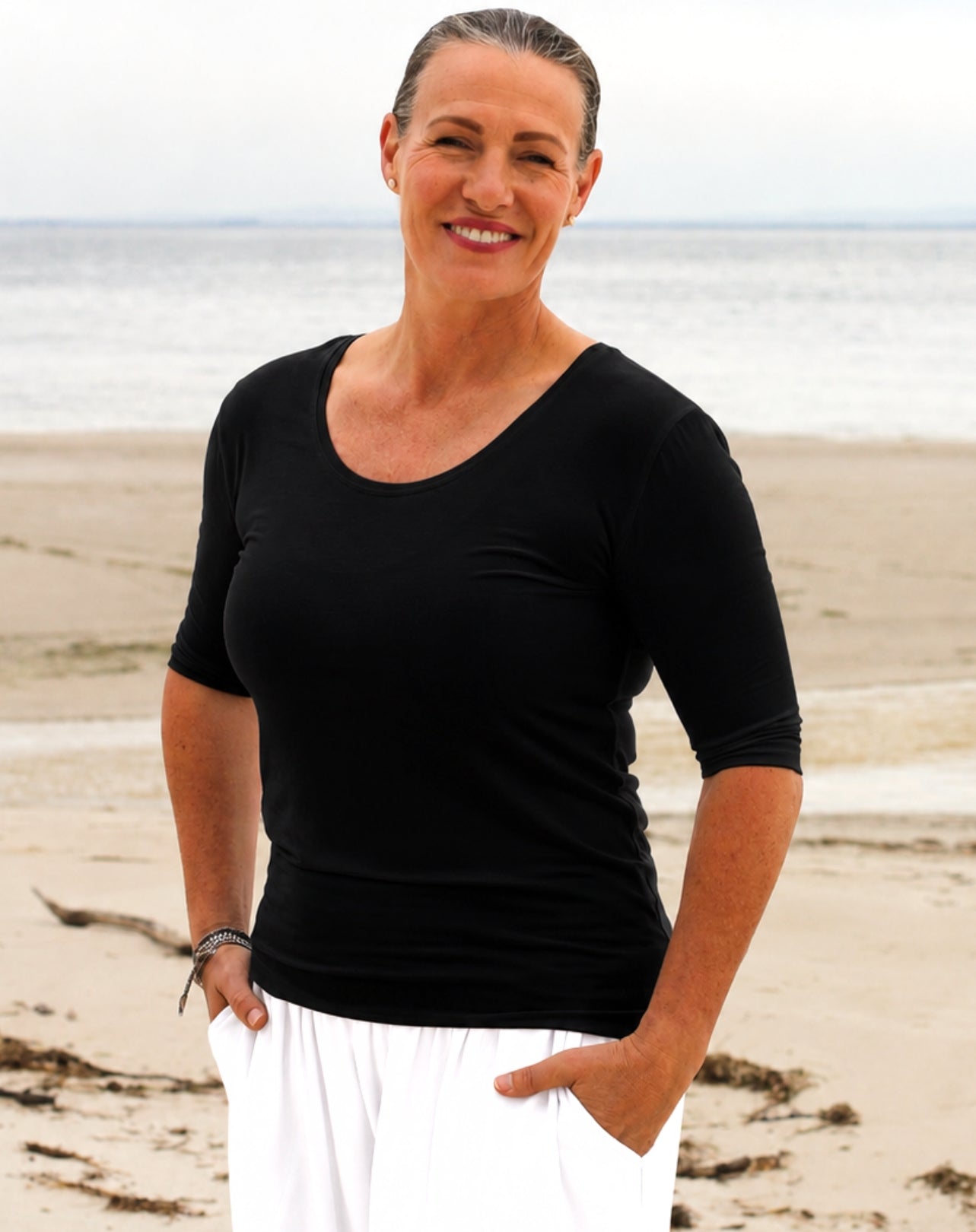A woman standing on a sandy beach wearing a black 3/4-sleeve scoop-neck top and white pants.