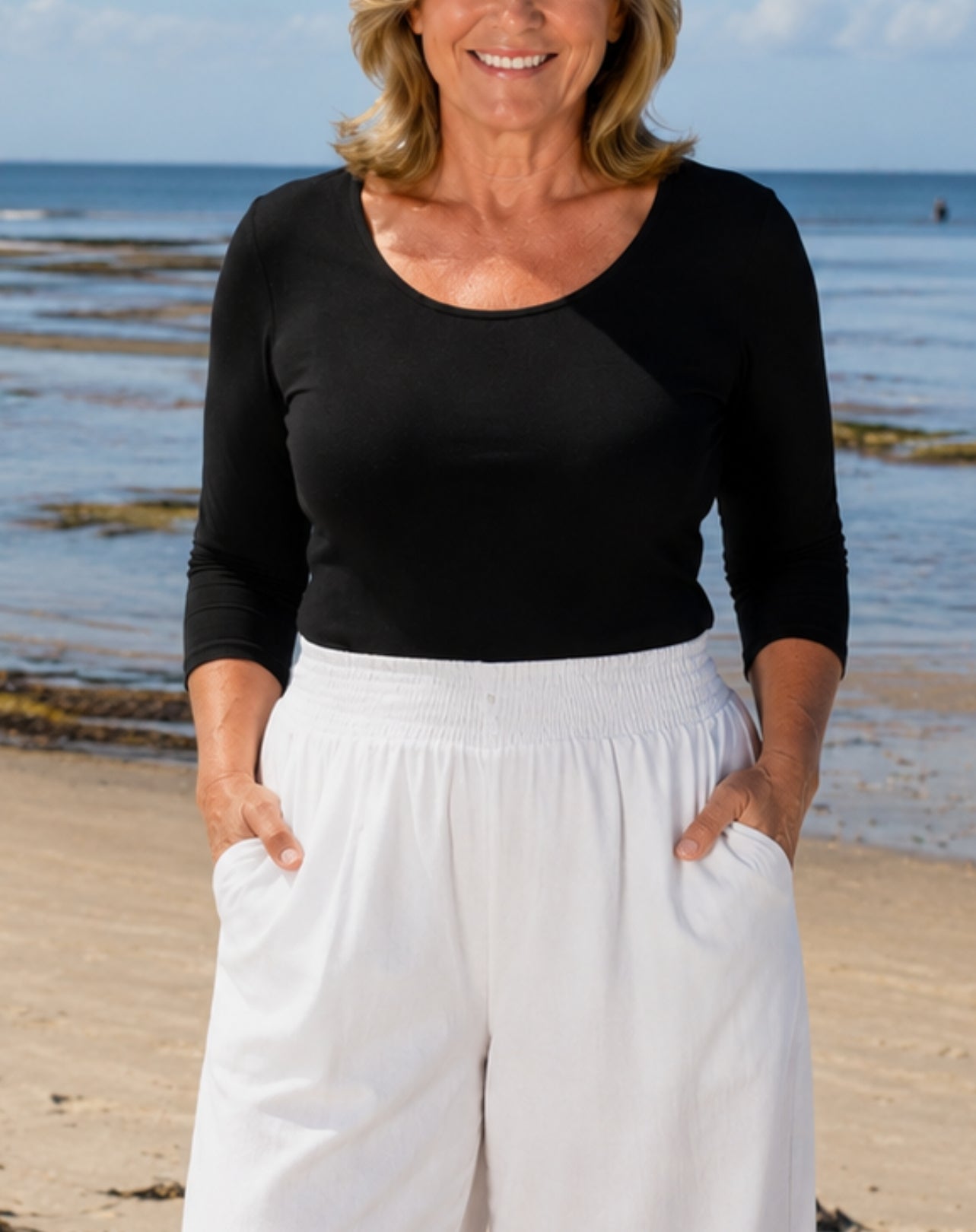 A woman standing on a beach wearing a black three-quarter-sleeve scoop-neck top and white wide-leg pants with side pockets.