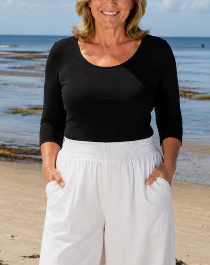 A woman standing on a beach wearing a black three-quarter-sleeve scoop-neck top and white wide-leg pants with side pockets.