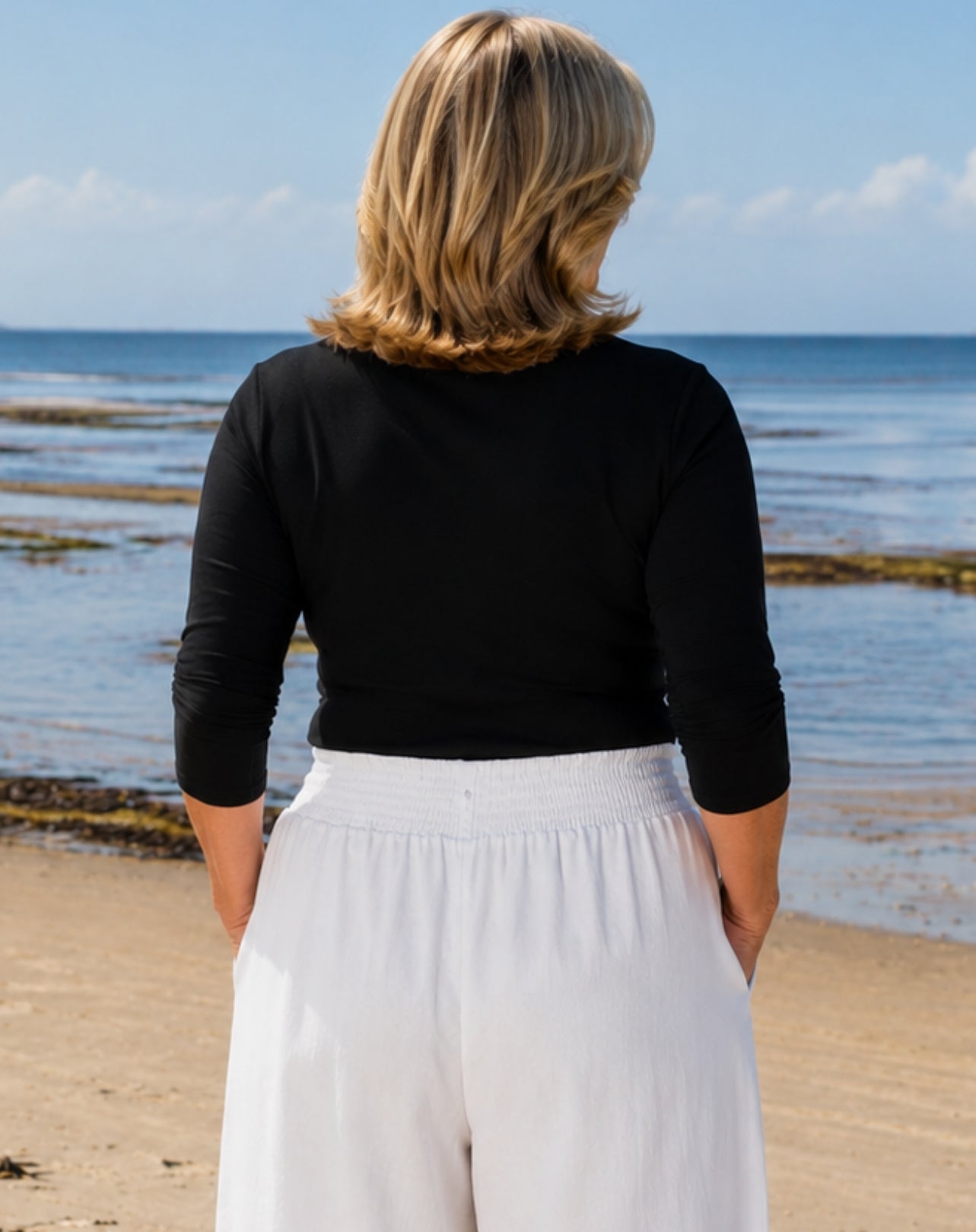 A woman viewed from behind standing on a sandy beach with the ocean and blue sky in the background. She is wearing a fitted black three-quarter-sleeve top and white wide-leg pants.