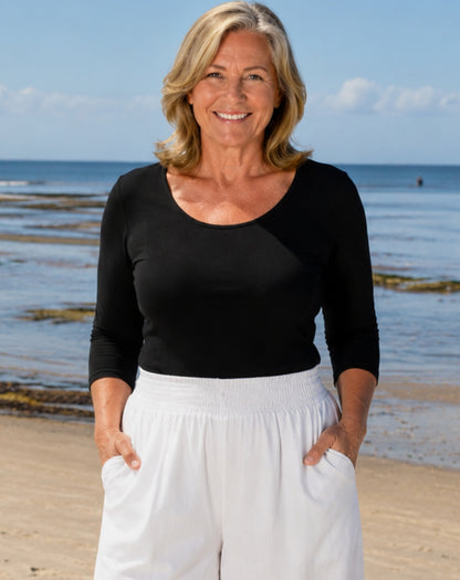 A smiling woman standing on a sandy beach with the ocean and blue sky behind her, wearing a black three-quarter-sleeve scoop-neck top and white drawstring pants.