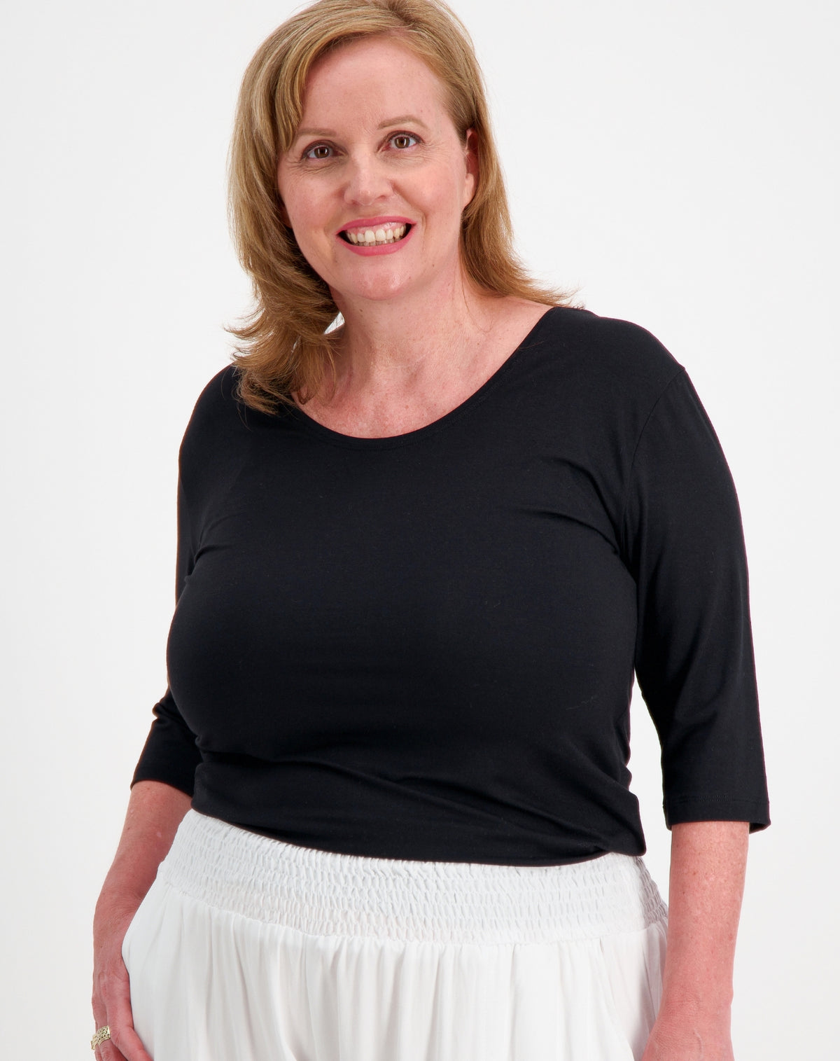 A smiling woman with blonde hair wearing a black three-quarter sleeve scoop-neck top and a white smocked pant, photographed against a plain white studio background.