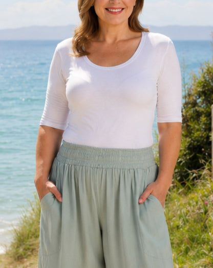 A woman wearing a white scoop-neck short-sleeve top and sage green wide-leg pants, standing outdoors with the ocean and grassy dunes in the background.