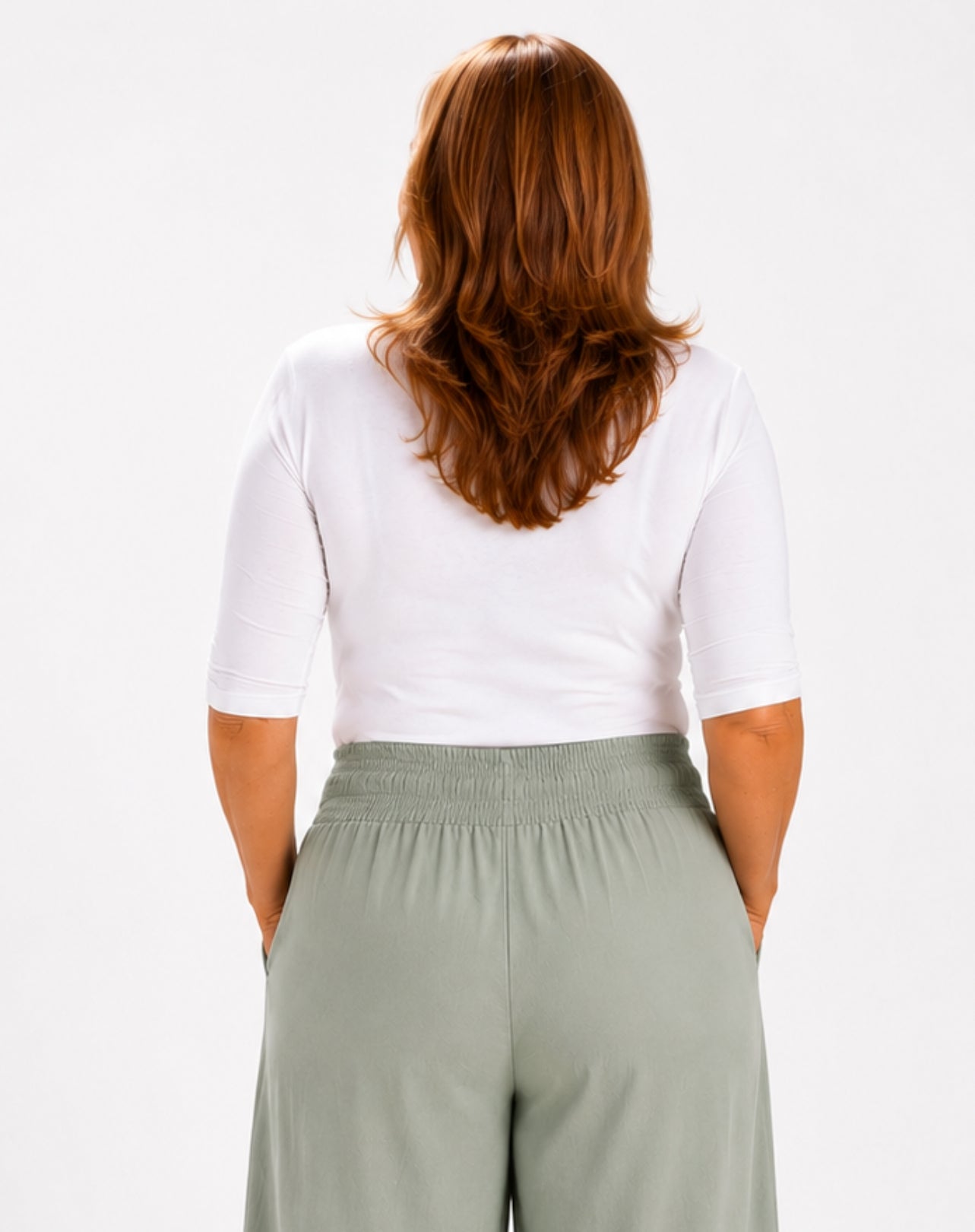 A woman viewed from the back wearing a white short-sleeve scoop tee and light mint green wide-leg pants against a plain light grey studio background.