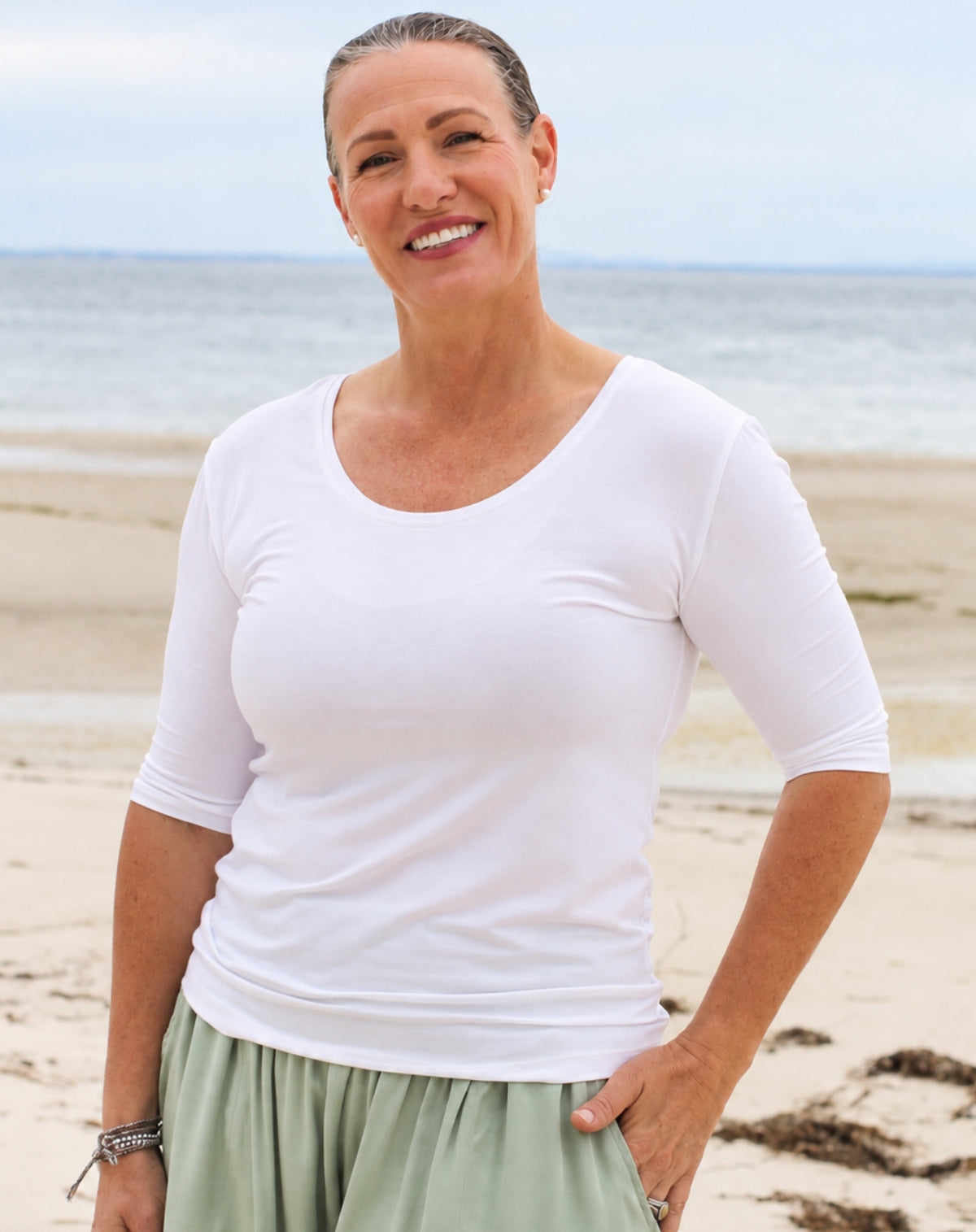A smiling woman standing on a sandy beach wearing a fitted white short-sleeve scoop-neck top and light sage green pants.