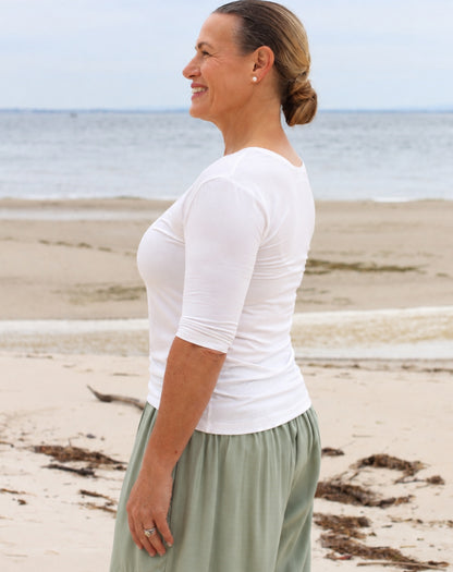 A woman standing on a sandy beach wearing a fitted white three-quarter-sleeve top and a light sage green skirt, shown in side profile with the ocean in the background.