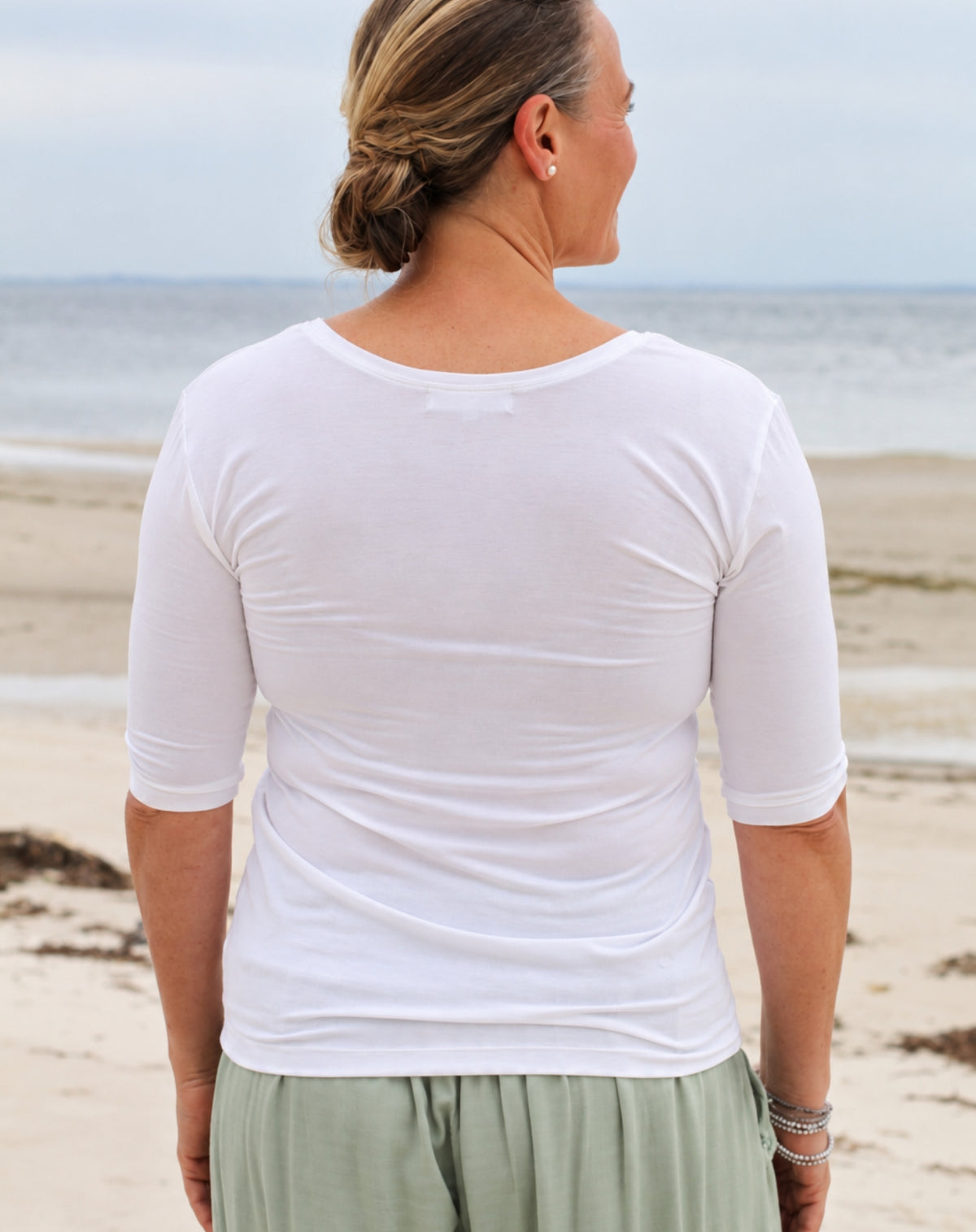 A woman standing on a beach, viewed from behind, wearing a fitted white short-sleeve top and light sage green skirt or pants.