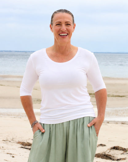 A smiling woman standing on a sandy beach wearing a white three-quarter sleeve scoop-neck top and mint green wide-leg pants.