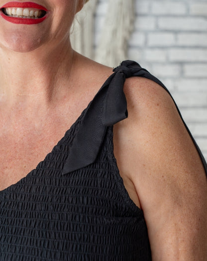 Woman wearing a black dress with a bow detail on her shoulder against a light brick wall.