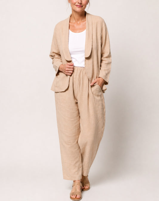 A woman modelling a natural linen-blend two-piece set consisting of a draped open-front blazer and matching comfort wide-leg pants, styled with a white tank top and tan sandals against a light grey studio background.