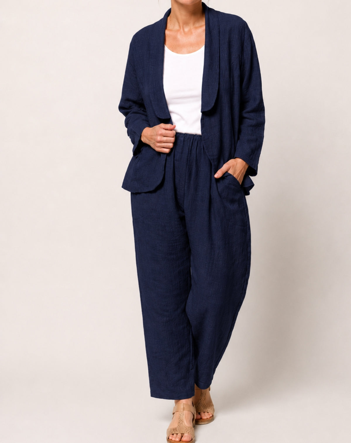 A woman modelling a navy blue two-piece linen set consisting of a light weight blazer and wide-leg pants, styled with a white tank top and beige sandals against a light beige studio background.