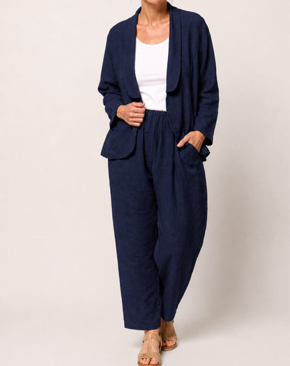 A woman modelling a navy blue two-piece linen set consisting of a light weight blazer and wide-leg pants, styled with a white tank top and beige sandals against a light beige studio background.