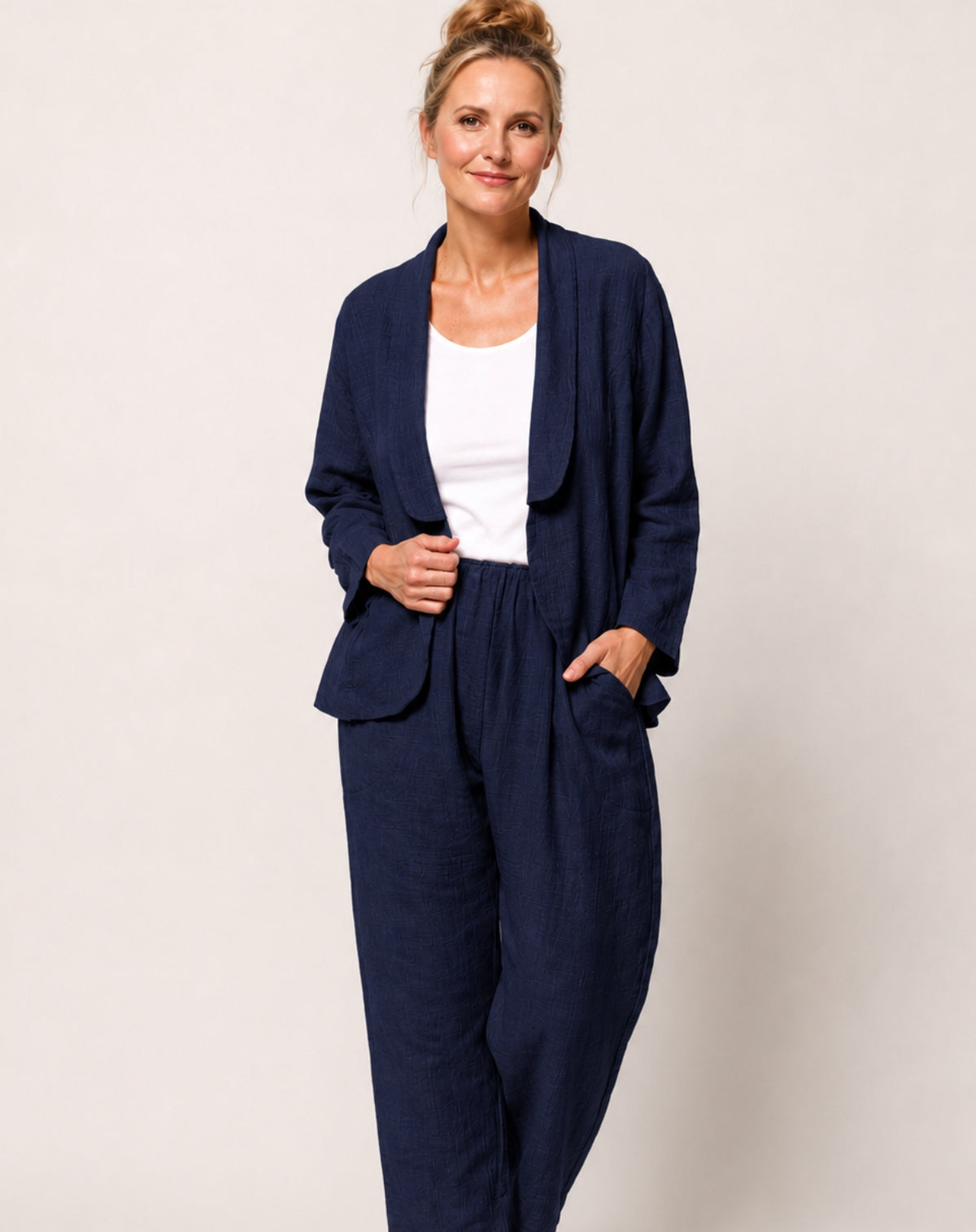 A woman modelling a navy blue cotton linen-blend two-piece set consisting of a long-sleeve open-front blazer jacket and matching relaxed trousers, worn over a white top, against a plain light grey studio background.