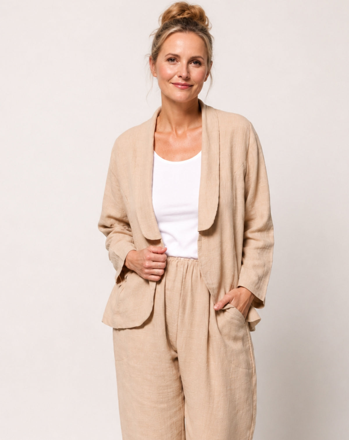 A woman modelling a natural linen two-piece set consisting of a cotton blazer and matching trousers, worn over a white scoop tee, against a plain light grey background.